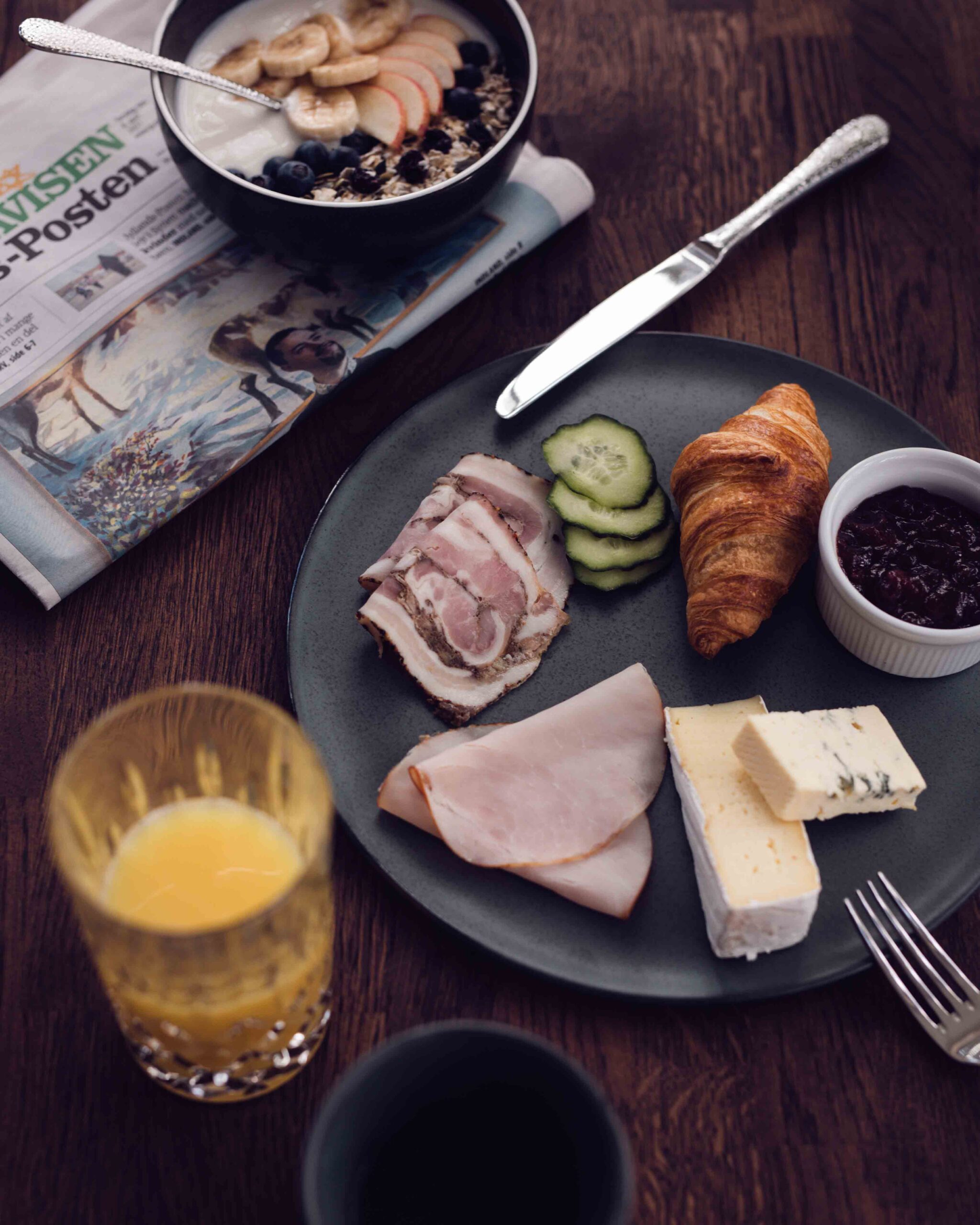 Et morgenmadsbord med en avis, et glas juice, en tallerken med hamburgerryg, ost, en croissant, en skål marmelade og en skål ved siden af med youghurt og frugt.
