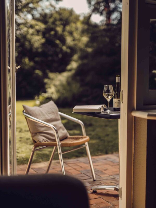 En åben dør ud til en terrasse med en stol og et bord, hvorpå der ligger en opslået bog og står et glas hvidvin.