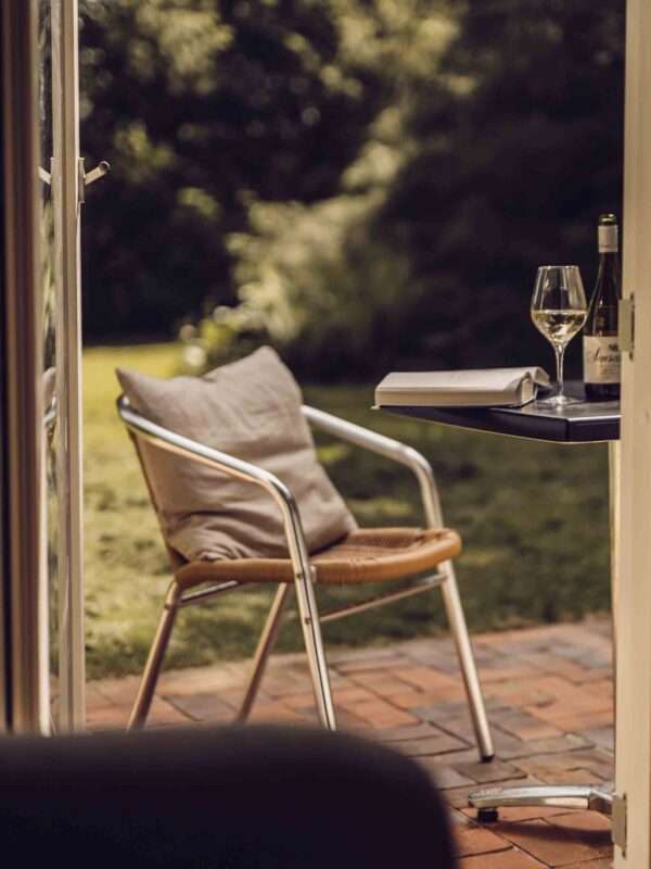 En åben dør ud til en terrasse med en stol og et bord, hvorpå der ligger en opslået bog og står et glas hvidvin.